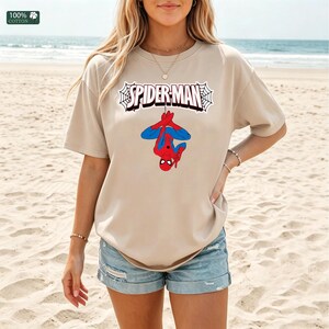 May include: Beige t-shirt with the Spider-Man logo and a graphic of Spider-Man hanging upside down. The text "SPIDER-MAN" is in a white web design. The shirt is made of 100% cotton.