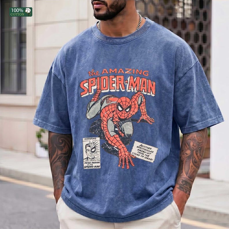 May include: A blue acid-wash t-shirt featuring a vintage-style graphic of Spider-Man in a crouching pose. The text reads "the AMAZING SPIDER-MAN" in orange, with a small comic book panel insert and a white box containing text about the character.