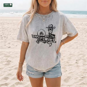 May include: A light gray short-sleeve t-shirt made of 100% cotton featuring a black line-art graphic of two cartoon characters, one resembling a space ranger and the other a cowboy, printed on the center of the chest with a crew neckline.