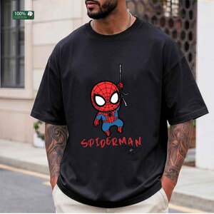 Op de afbeelding: Zwart T-shirt met een cartoon Spider-Man ontwerp. De afbeelding toont een rood en blauw Spider-Man karakter dat aan een web hangt, met het woord "SPIDERMAN" in het rood eronder. Het shirt is gemaakt van 100% katoen.