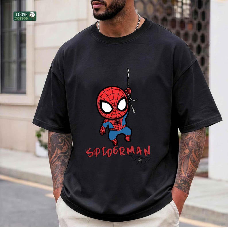Op de afbeelding: Zwart T-shirt met een cartoon Spider-Man ontwerp. De afbeelding toont een rood en blauw Spider-Man karakter dat aan een web hangt, met het woord "SPIDERMAN" in het rood eronder. Het shirt is gemaakt van 100% katoen.