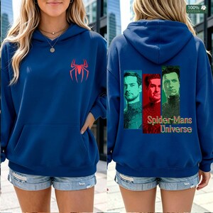 May include: A blue hooded sweatshirt featuring a red spider logo on the front chest. The back displays three vertical rectangular portraits of men in superhero costumes with the text Spider-Mans Universe in a stylized font. The fabric is 100% cotton.