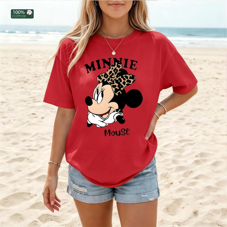 May include: Red t-shirt featuring a Minnie Mouse graphic. The graphic includes the text "MINNIE" above the character's head and "Mouse" below. The shirt is made of 100% cotton.