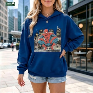 May include: A dark blue hooded sweatshirt featuring a graphic print of the comic book character Spider-Man crouching in a city setting. The hoodie includes a front kangaroo pocket, long sleeves, and a drawstring hood, made from 100% cotton fabric.