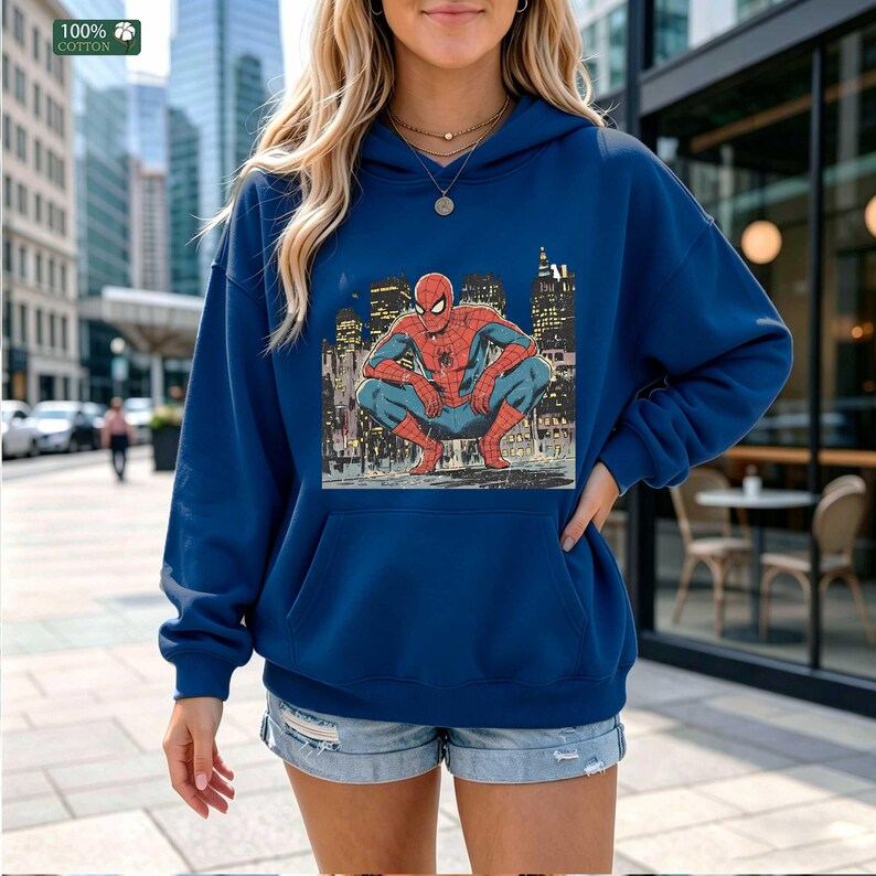 May include: A dark blue hooded sweatshirt featuring a graphic print of the comic book character Spider-Man crouching in a city setting. The hoodie includes a front kangaroo pocket, long sleeves, and a drawstring hood, made from 100% cotton fabric.