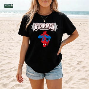 May include: Black t-shirt with the Spider-Man logo and a graphic of Spider-Man in a red and blue suit. The text "SPIDER-MAN" is in white, outlined in black, and set against a white web design. The shirt is made of 100% cotton.