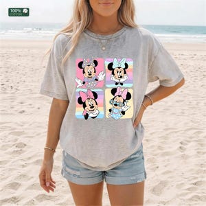May include: A heather gray short-sleeve t-shirt featuring a graphic print of four squares, each containing a different illustration of Minnie Mouse in various poses and outfits, with a small 100% cotton label visible in the upper left corner.