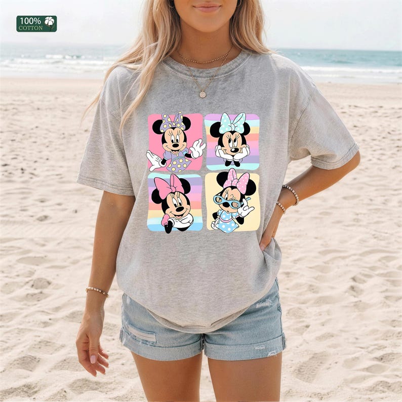 May include: A heather gray short-sleeve t-shirt featuring a graphic print of four squares, each containing a different illustration of Minnie Mouse in various poses and outfits, with a small 100% cotton label visible in the upper left corner.