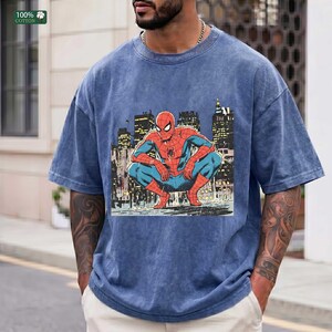 May include: A blue acid-wash t-shirt featuring a graphic print of the comic book character Spider-Man crouching in a city setting. The character is depicted in a red and blue suit against a dark, detailed urban skyline background with lit windows.