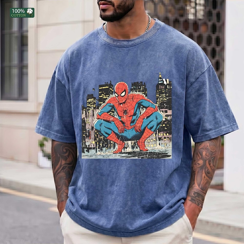 May include: A blue acid-wash t-shirt featuring a graphic print of the comic book character Spider-Man crouching in a city setting. The character is depicted in a red and blue suit against a dark, detailed urban skyline background with lit windows.