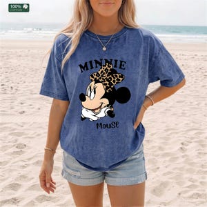 May include: Blue t-shirt featuring a Minnie Mouse graphic. The graphic includes Minnie's face, a leopard print bow, and the text "MINNIE MOUSE". The shirt is made of 100% cotton.