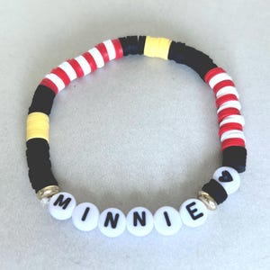 Puede incluir: Una pulsera de cuentas con el nombre "MINNIE" escrito con cuentas de letras blancas. La pulsera presenta cuentas negras, rojas, blancas y amarillas. La pulsera tiene una cuenta en forma de coraz&oacute;n y detalles dorados.
