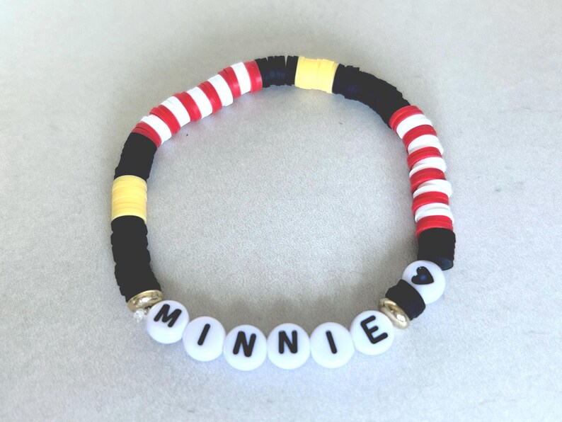 Puede incluir: Una pulsera de cuentas con el nombre "MINNIE" escrito con cuentas de letras blancas. La pulsera presenta cuentas negras, rojas, blancas y amarillas. La pulsera tiene una cuenta en forma de coraz&oacute;n y detalles dorados.