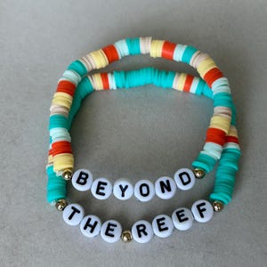 May include: Two beaded bracelets with the text "BEYOND THE REEF". The bracelets are made of colourful, flat, round beads in shades of turquoise, orange, yellow, and white. Gold-coloured beads separate the text. The bracelets are on a grey surface.