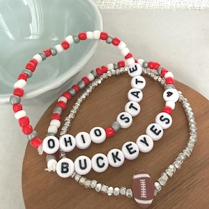 May include: Three beaded bracelets featuring red, white, and silver beads. One bracelet displays the text OHIO STATE with a heart, another features the text BUCKEYES, and the third includes a brown and white football-shaped bead charm.