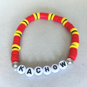 Op de afbeelding: Een armband van rode, gele en zwarte kralen met het woord "KACHOW" gespeld in witte letterkralen. De armband is gemaakt van platte, ronde kralen en heeft zilverkleurige eindkappen.