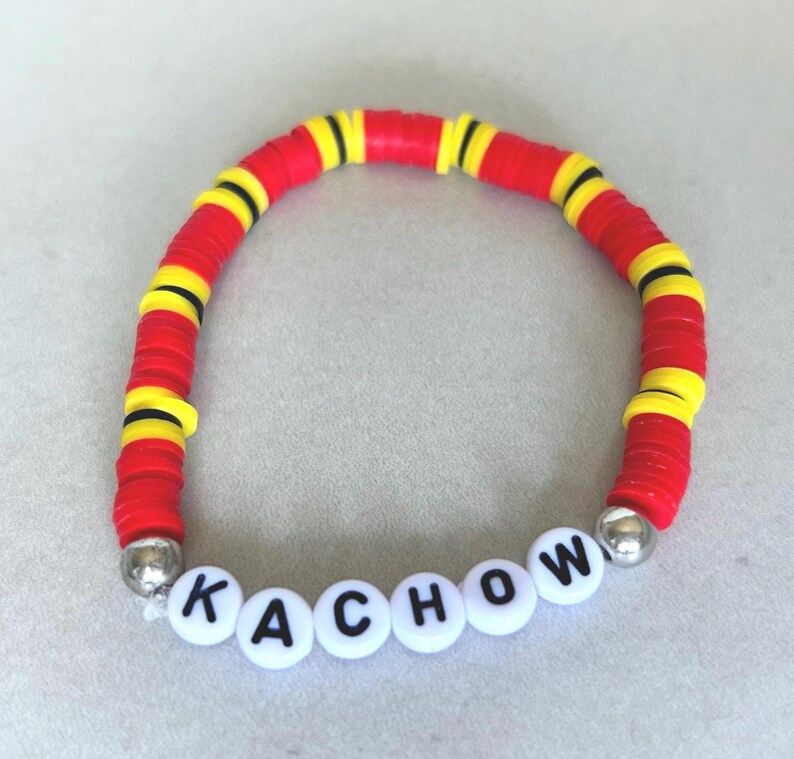 Op de afbeelding: Een armband van rode, gele en zwarte kralen met het woord "KACHOW" gespeld in witte letterkralen. De armband is gemaakt van platte, ronde kralen en heeft zilverkleurige eindkappen.