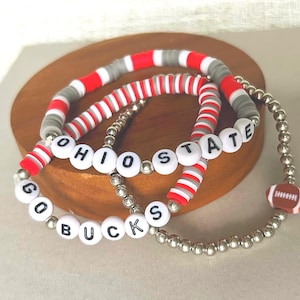 Peut inclure: Trois bracelets avec des perles rouges, blanches et grises. L'un des bracelets épelle "OHIO STATE" avec des perles de lettres blanches. Un autre dit "GO BUCKS". Un charm de football est attaché. Les bracelets sont sur une surface en bois.