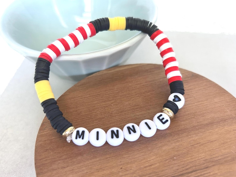 Puede incluir: Una colorida pulsera de cuentas con el nombre "Minnie" escrito con cuentas de letras blancas. La pulsera presenta cuentas a rayas rojas y blancas, cuentas negras y amarillas y una cuenta en forma de coraz&oacute;n.