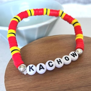 Op de afbeelding: Een kleurrijke armband met rode, gele en zwarte kralen, met het woord "KACHOW" gespeld met witte letterkralen. De armband ligt op een houten oppervlak.