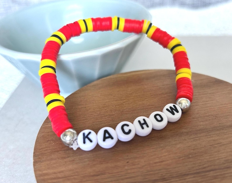 Op de afbeelding: Een kleurrijke armband met rode, gele en zwarte kralen, met het woord "KACHOW" gespeld met witte letterkralen. De armband ligt op een houten oppervlak.