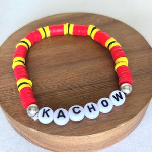 Op de afbeelding: Een rode, gele en zwarte kralenarmband met het woord "KACHOW" gespeld in witte letterkralen. De armband is gemaakt van platte, schijfvormige kralen en heeft zilverkleurige eindkappen.