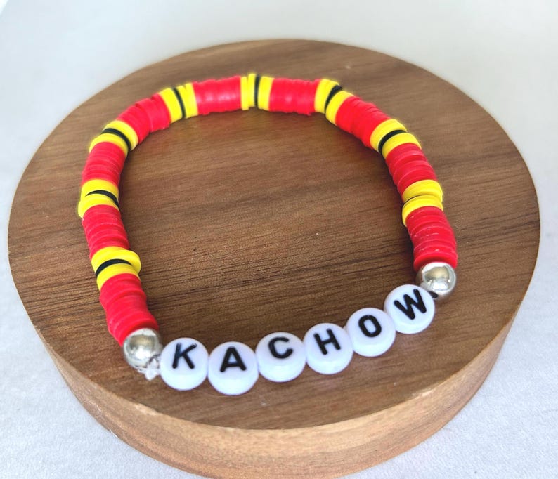 Op de afbeelding: Een rode, gele en zwarte kralenarmband met het woord "KACHOW" gespeld in witte letterkralen. De armband is gemaakt van platte, schijfvormige kralen en heeft zilverkleurige eindkappen.