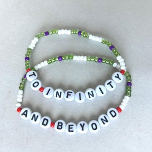 May include: Two beaded bracelets. The top bracelet says "TO INFINITY" and the bottom bracelet says "AND BEYOND" in white letter beads. The bracelets have white, green, purple, and red beads.