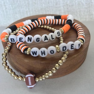 Könnte beinhalten: Drei Armbänder mit Perlen, auf denen die Wörter "BENGALS" und "WHO DEY" mit weißen Buchstabenperlen geschrieben sind. Die Armbänder haben orange, schwarze, weiße und goldene Perlen mit einem Football-Anhänger. Die Armbänder werden auf einer Holzoberfläche präsentiert.