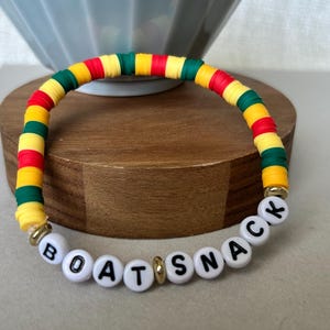 May include: A colorful bracelet with the word "BOATSNAK" spelled out in white letter beads. The bracelet features a pattern of red, yellow, green, and gold beads. The bracelet is resting on a wooden surface.
