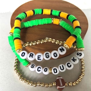 Può includere: Tre braccialetti impilati. I due braccialetti superiori sono realizzati con perline piatte verdi e gialle con accenti neri. Il braccialetto inferiore ha perline dorate e perline con lettere bianche che recitano "OREGON SCO DUCKS". È attaccato un ciondolo a forma di pallone da football.