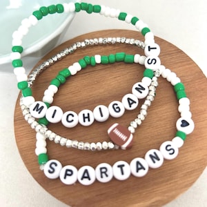Peut inclure: Trois bracelets de perles composés de perles rondes blanches et vertes, de perles intercalaires métalliques argentées et de perles lettres blanches formant MICHIGAN ST et SPARTANS. Un bracelet inclut une perle ballon de football et un cœur noir.