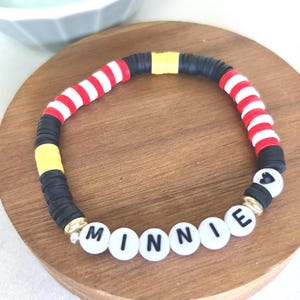 Puede incluir: Una colorida pulsera de cuentas con el nombre "MINNIE" escrito con cuentas de letras blancas. La pulsera presenta cuentas a rayas rojas y blancas, cuentas negras y cuentas amarillas, con detalles dorados.