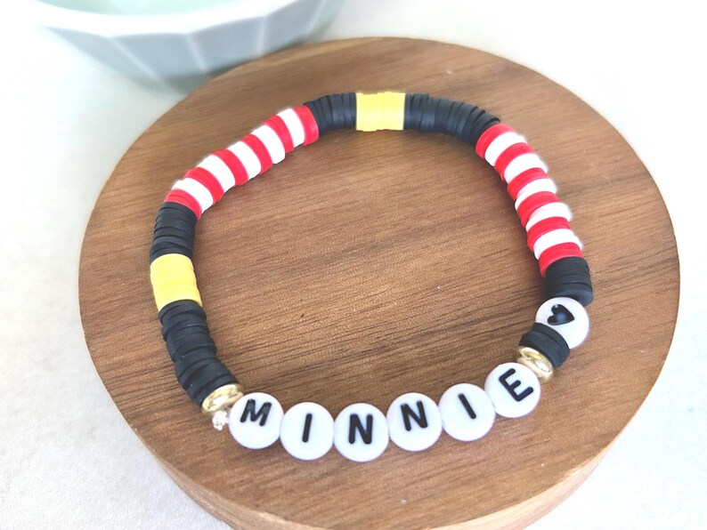 Puede incluir: Una colorida pulsera de cuentas con el nombre "MINNIE" escrito con cuentas de letras blancas. La pulsera presenta cuentas a rayas rojas y blancas, cuentas negras y cuentas amarillas, con detalles dorados.