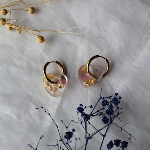 May include: A pair of gold hoop earrings featuring clear, heart-shaped resin charms. Each charm is embedded with small, shimmering gold flakes and subtle pink accents, suspended from a polished gold-tone metal hoop.