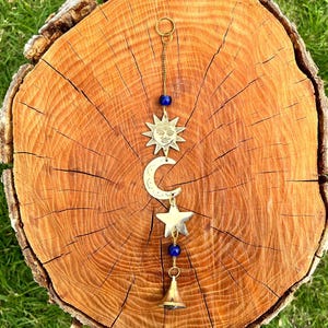 Op de afbeelding: Een windgong met een hemels thema met een zon, maan en ster ontwerp. De metalen ornamenten zijn opgehangen aan een gouden ketting en versierd met blauwe kralen. De windgong heeft een kleine bel aan de onderkant. De achtergrond is een houten boomstronk.
