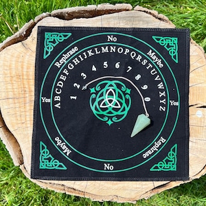 May include: A black divination board with a green Celtic knot design and lettering. The board has the words "Yes", "No", and "Maybe" and numbers 1-9. A green crystal pendulum hangs over the board.