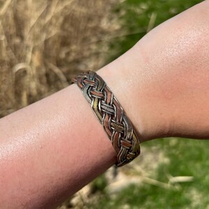 May include: A braided metal bracelet in silver, copper, and gold tones. The bracelet is worn on a person's arm, showcasing the intricate woven design. The background features blurred natural elements.