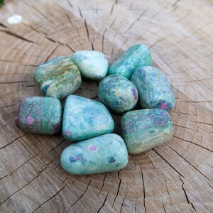 Può includere: Un gruppo di pietre burattate nei toni del verde acqua, verde e blu, con macchie rosa e rosse. Le pietre sono lisce e arrotondate, appoggiate su una superficie di legno stagionato. Queste pietre preziose sono ideali per la meditazione o l'esposizione.