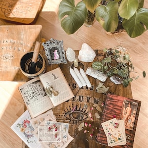 Könnte beinhalten: Ein hölzerner Spirit-Board-Tisch mit einer schwarzen Klangschale samt Holzschlägel, einem verzierten silbernen Rahmen, drei weißen Kerzen, rohen Quarzkristallen, einem Buch mit okkulten Diagrammen, Tarotkarten, getrockneten Rosenknospen und einer grünen Topfpflanze.