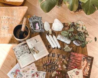 Puede incluir: Una mesa de madera tipo tablero de espiritismo con un cuenco tibetano negro y mazo de madera, un marco plateado ornamentado, tres velas blancas, cristales de cuarzo, un libro con diagramas ocultos, cartas de tarot, capullos de rosa secos y una planta verde.