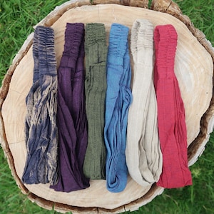 May include: Six colorful headbands in shades of blue, purple, green, blue, beige, and red are arranged on a wooden surface. The headbands are made of a soft, textured fabric.