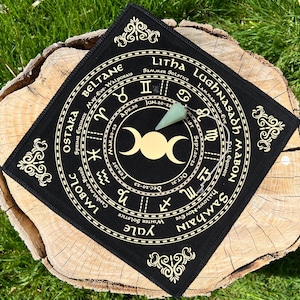 May include: A black divination mat with gold-colored text and symbols, including zodiac signs and phases of the moon. A green crystal pendulum rests on the mat. The mat is square and sits on a wooden surface.
