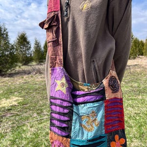 Peut inclure: Un sac à bandoulière patchwork avec une longue sangle. Le sac présente une variété de couleurs et de motifs, notamment du violet, du sarcelle et de l'orange. Il comporte une étoile et un symbole Om. Le sac est fait d'un tissu doux et texturé.