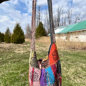 Può includere: Una colorata borsa a tracolla patchwork con una lunga tracolla. La borsa presenta varie toppe di tessuto in tonalità di rosso, arancione, blu, viola e verde, decorate con disegni a fantasia. La borsa è sospesa su uno sfondo di un campo erboso e un cielo parzialmente nuvoloso.