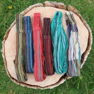 May include: Seven colourful headbands with elasticated backs, arranged on a wooden tree stump against green grass. The headbands display striped patterns in blue, red, green, and brown. The headbands are made of fabric.