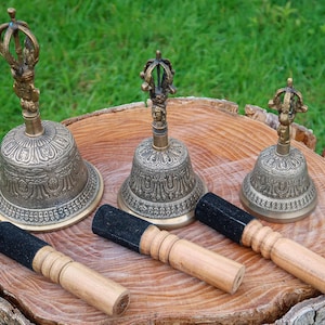 May include: Three brass bells with ornate designs and wooden strikers with black handles are displayed on a tree stump. The bells vary in size, with the largest bell on the left. The background is green grass.