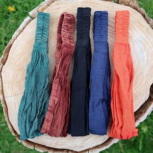 May include: Five headbands in teal, rust, black, blue, and orange are displayed on a wooden surface. The headbands have a ruched texture and are arranged in a row. The background is green grass.