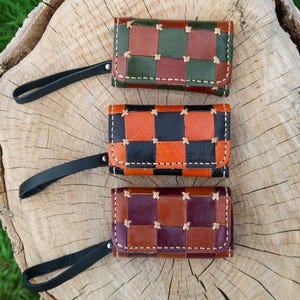 May include: Three handmade leather key wallets with a checkered pattern in various colours: green, brown, orange, black, and purple. Each wallet has a black wrist strap. The wallets are displayed on a wooden surface.