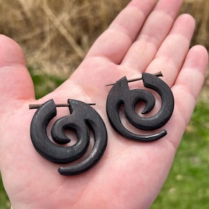 May include: Two dark brown wooden spiral earrings. Each earring has a swirling design and a straight post. The earrings are shown on an open palm, with a blurred background of grass and foliage. The earrings are a unique accessory.
